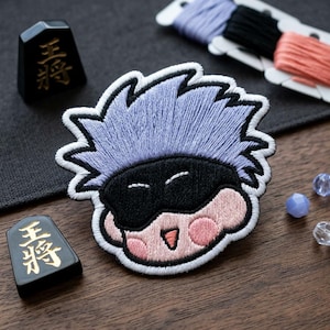 May include: Embroidered patch featuring a cartoon character with light blue hair, a black mask, and pink cheeks. The patch is surrounded by a white border. Two black game pieces with gold Japanese characters are also visible.