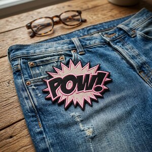 May include: Blue denim jeans with a distressed appearance, displaying a "POW!" patch. The patch features a pink and black design with a starburst background. Tortoiseshell spectacles are on a wooden surface.