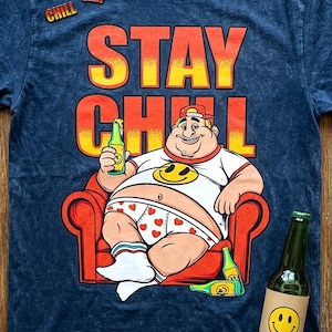 May include: Blue t-shirt with a graphic of a cartoon man relaxing in a red chair, holding a beer. The shirt has the words "STAY CHILL" in orange and yellow. Additional details include a smiley face and a small red chair graphic.