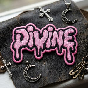 May include: A pink embroidered patch with the word "DIVINE" in a dripping font, outlined in black, rests on a textured brown surface. Silver necklaces with cross and crescent moon pendants surround the patch, along with safety pin charms.