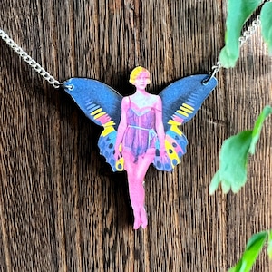 May include: A silver chain necklace with a cutout of a fairy with blue wings, a pink dress, and yellow accents. The fairy is standing on a wooden surface.