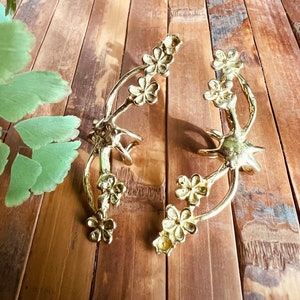 May include: Two gold-toned metal ear cuffs with a floral design. Each cuff features a delicate branch with multiple flower-shaped accents.
