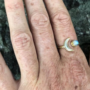 May include: A gold crescent moon ring with a white moonstone accent. The ring is worn on a person's finger.