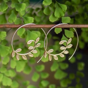 May include: A pair of gold hoop earrings with a delicate leaf design. The earrings are hanging from a brown metal rod.