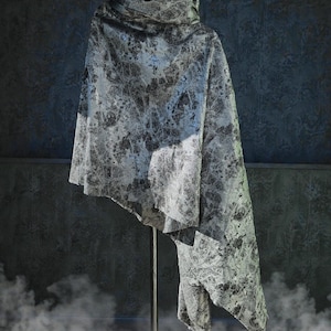 May include: A draped, abstract-patterned cape in shades of gray and black hangs from a metal stand. The fabric has a textured appearance with a marbled design. The cape's design includes a high, cowl-like collar.