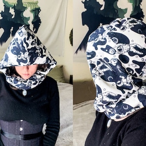 May include: A white and black hooded scarf with a space-themed print featuring astronauts and stars. The scarf is made of a soft, comfortable fabric and can be worn as a neck warmer or a hood.