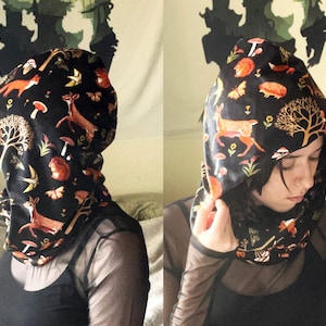 May include: A black hooded cowl with a colorful woodland print featuring deer, mushrooms, trees, and birds. The cowl is made of a soft, stretchy fabric and is perfect for keeping warm in the winter.