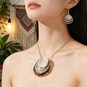 May include: A gold-toned necklace and matching earrings set. The necklace features a pendant with three layered, irregularly shaped elements in white, silver, and burgundy. The earrings have a similar design. The jewelry is worn against a skin tone background.