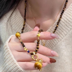May include: A necklace featuring brown tiger eye beads and a gold-toned clasp. The piece includes a gold-toned pendant shaped like a gourd, accented with a small blue bead and a gold-toned spacer. The necklace is strung on a thin cord.