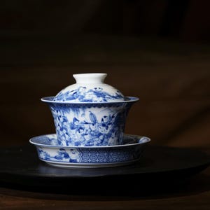 以下が含まれることがあります： 青と白の磁器のティーセット。蓋付きの蓋碗と、それに合うソーサーが含まれています。蓋碗とソーサーは、人物や模様の詳細なイラストで装飾されています。ティーセットは暗い表面に置かれています。