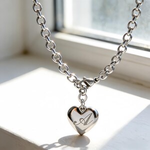 Op de afbeelding: Zilverkleurige ketting met een hartvormige hanger. De hanger heeft een gegraveerde sierlijke letter "A". De ketting heeft ronde schakels en een karabijnsluiting. De ketting wordt getoond op een witte ondergrond.