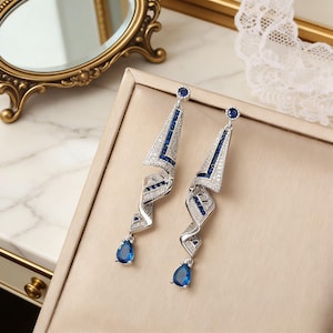 Puede incluir: Un par de pendientes colgantes plateados con un diseño geométrico. Los pendientes presentan una combinación de piedras preciosas claras y azules, con una piedra azul en forma de lágrima en la parte inferior. Los pendientes se exhiben sobre una superficie de tono neutro.