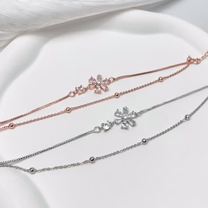 Peut inclure: Deux bracelets délicats avec des motifs floraux. L'un est en or rose, l'autre en argent. Chaque bracelet présente une pièce maîtresse en forme de fleur avec des cristaux transparents. Les bracelets ont de petites perles rondes et une chaîne délicate.