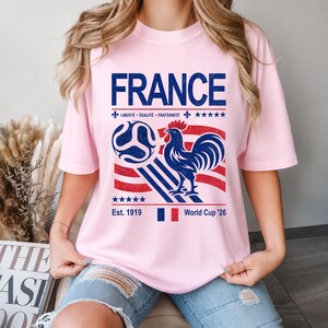 May include: A light pink t-shirt featuring a graphic design with the word "FRANCE" in blue, a soccer ball, a rooster, and the text "Libert&eacute;, &Eacute;galit&eacute;, Fraternit&eacute;". The shirt also includes the text "Est. 1919" and "World Cup '26".