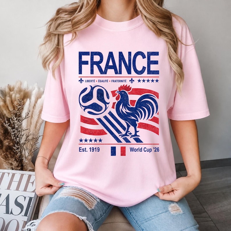 May include: A light pink t-shirt featuring a graphic design with the word "FRANCE" in blue, a soccer ball, a rooster, and the text "Libert&eacute;, &Eacute;galit&eacute;, Fraternit&eacute;". The shirt also includes the text "Est. 1919" and "World Cup '26".