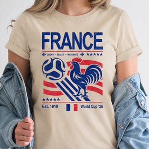 May include: Beige t-shirt with a blue and red graphic featuring the word "FRANCE", a soccer ball, a rooster, and the text "Est. 1919" and "World Cup '26". The shirt also has the French motto "Libert&eacute;, &Eacute;galit&eacute;, Fraternit&eacute;".