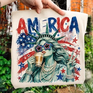 Könnte beinhalten: Eine cremefarbene Tragetasche mit einer grafischen Illustration der Freiheitsstatue, die sternförmige Sonnenbrillen trägt, eine Kaffeetasse hält und ein Friedenszeichen macht. Das Design enthält das Wort AMERICA, Feuerwerk und Sterne.