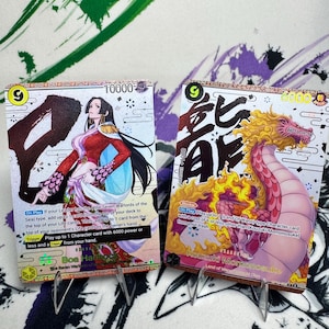 Może przedstawiać: Dwie karty kolekcjonerskie z postaciami z anime. Jedna karta przedstawia kobietę w czerwono-złotym stroju z napisem "Boa Hancock". Druga karta przedstawia stworzenie podobne do smoka z napisem "Kouzuki Momonosuke". Obie karty mają poziom mocy i instrukcje gry.