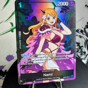 May include: A holographic One Piece trading card featuring the character Nami with orange hair and horns, wearing a purple and white outfit. The card displays the text On Play, Land of Wano, and Counter +1000 against a dark, patterned background.