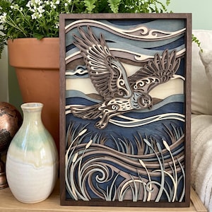 May include: A layered wooden art piece depicting an owl in flight. The artwork features a dark brown frame, with the owl in shades of brown and tan against a blue and white background. The bottom of the piece shows reeds.