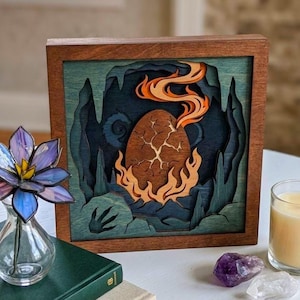 Pode incluir: Uma caixa de sombra quadrada de madeira com um design recortado em camadas de um ovo rachado cercado por chamas laranja dentro de uma caverna azul escura. A obra de arte é emoldurada em madeira de tom médio e exibe profundidade complexa.