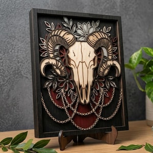 May include: A framed, layered wooden art piece featuring a ram skull design. The artwork is primarily in shades of brown, cream, and black, with floral and chain accents. The piece is displayed on a wooden easel.