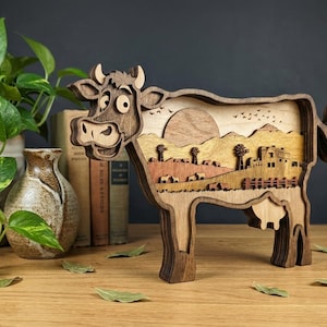Op de afbeelding: Houten kunstwerk in de vorm van een koe met een gelaagd ontwerp. De koe heeft een lachend gezicht en een gedetailleerde landschapsbinnenkant, inclusief een zon, bergen en boerderijgebouwen. Het stuk is gemaakt van verschillende bruintinten.