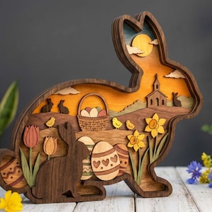 Puede incluir: Decoración de conejo de Pascua de madera con diseño en capas. Presenta una escena de puesta de sol con una iglesia, conejos y una cesta de huevos. Incluye tulipanes, narcisos y huevos de Pascua decorados. Un acento primaveral.