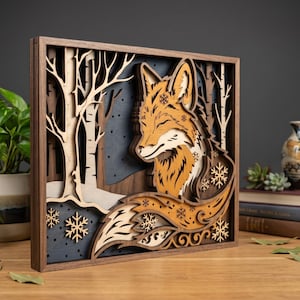 May include: A multi-layered wooden artwork depicting a fox in a winter forest. The fox is orange and white, with snowflake patterns. The background shows trees and a dark blue sky, framed in dark brown wood.