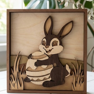Puede incluir: Arte de pared de Pascua de madera. Un conejo marrón sonriente sostiene un huevo decorativo. La obra de arte está enmarcada en un marco cuadrado de madera marrón, con recortes en capas de hierba y un fondo.