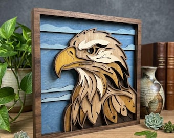 May include: A square wooden wall art piece featuring a multi-layered laser-cut eagle head profile. The eagle is crafted from various shades of natural wood, set against a blue background with horizontal wave patterns, all encased in a dark frame.
