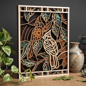 May include: A multi-layered wooden wall art piece featuring a bird perched on a branch with leaves. The artwork uses shades of brown, orange, and teal, with a light wood frame. The design includes a sun-like element and intricate leaf patterns.