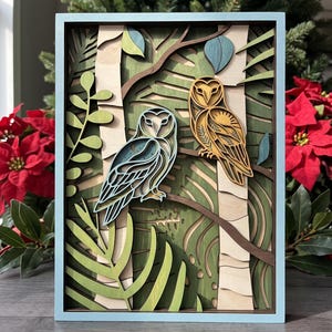 May include: A multi-layered wooden wall art piece featuring two owls perched on branches within a forest setting. The artwork displays a light blue frame, birch-colored tree trunks, and various shades of green foliage, with one owl in blue and one in gold.