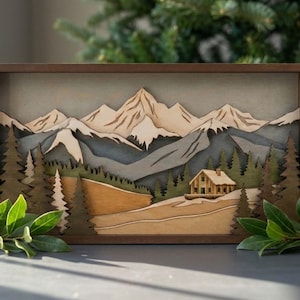 May include: A wooden mountain landscape art piece. The artwork features a cabin, trees, and snow-capped mountains in a layered design. The color palette includes shades of brown, green, gray, and beige, with a dark brown frame.