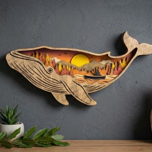 Puede incluir: Arte de pared de madera con forma de ballena con un diseño en capas que representa una escena de puesta de sol con árboles, montañas y un barco. La ballena es de madera clara y cuelga de una pared gris oscuro.