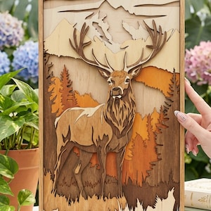 May include: A wooden layered art piece featuring a majestic deer in a forest scene. The artwork uses various shades of brown and orange to depict the deer, trees, and mountains. The piece is framed in a light-colored wood.
