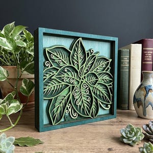 May include: A layered wooden wall art piece featuring a detailed green leaf design within a teal frame. The artwork showcases intricate cut-out patterns and varying shades of green, creating a three-dimensional effect. The piece is displayed on a wooden surface.