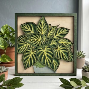 May include: A layered wooden art piece featuring a detailed depiction of a houseplant. The leaves are a mix of green and yellow, set against a light wood background, framed in a dark green square. The plant is in a light blue pot.