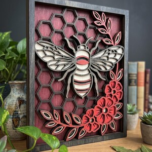 Può includere: Opera d'arte in legno a strati con un design a forma di ape. L'opera comprende un'ape, un favo, fiori e foglie, tutti nei toni del rosso, bianco e nero, all'interno di una cornice in legno scuro. Un oggetto decorativo.