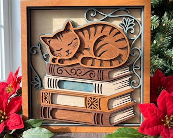 May include: A multi-layered wooden wall art piece featuring a sleeping orange tabby cat resting on a stack of four books. The design includes intricate scrollwork accents in a light blue hue, all encased within a warm-toned wooden square frame.