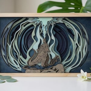 Peut inclure: Une œuvre d'art murale en bois multicouche représentant une figurine centrale de Godzilla émettant un rayon d'énergie bleu clair. Le design utilise des nuances de bleu marine, de sarcelle et de vert clair dans un style découpé, encadré de bois clair.