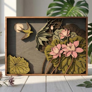 Puede incluir: Arte mural de madera con un diseño en capas de un pájaro en vuelo, flores de loto y hojas. La obra de arte está enmarcada en madera oscura, con un fondo gris neutro. Las flores son rosas y las hojas son verdes.
