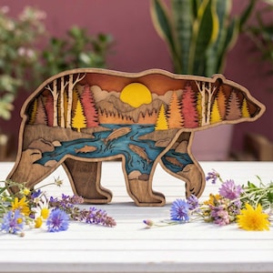 May include: A wooden bear-shaped art piece with a layered design. The interior depicts a landscape with a river, fish, trees, and a sunset. The bear is brown, and the landscape features shades of orange, yellow, and blue. Flowers surround the base.