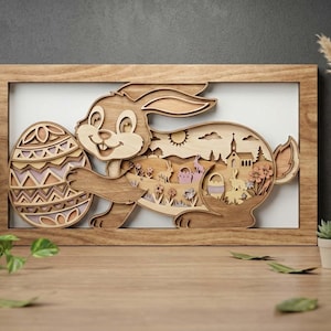 Puede incluir: Obra de arte de conejo de Pascua de madera con un diseño en capas. El conejo sostiene un huevo decorativo con un patrón detallado. La escena interior presenta una iglesia, árboles y elementos de Pascua. La obra está enmarcada en un marco de madera natural.