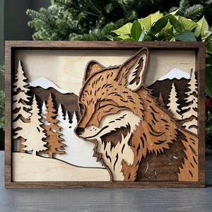 Puede incluir: Obra de arte enmarcada en madera que presenta un diseño en capas de un zorro en un paisaje invernal. El zorro está representado en tonos marrones y crema, con una expresión serena. El fondo incluye montañas nevadas y pinos.
