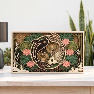 May include: A rectangular multi-layered wooden wall art piece featuring two koi fish swimming in a circular pattern. The design includes pink lotus flowers, green lily pads, and bamboo stalks set against a tan background with wave patterns.