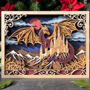 May include: A multi-layered wooden shadow box art piece featuring a purple dragon with spread wings perched atop a tan castle. The scene includes snow-capped brown mountains, a red moon, and blue clouds against a dark sky, all framed by ornate scrollwork.