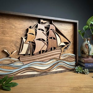 May include: Wooden layered art piece depicting a sailing ship on the ocean. The artwork features a dark brown frame, with the ship and waves in shades of brown, tan, and blue. A small wooden bird is also included.