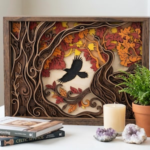 May include: A layered wooden art piece depicting a black bird in flight through a forest scene with autumn leaves in shades of red, orange, and yellow. The artwork is framed in a dark brown wood and sits on a white surface. A candle, crystals, and books are also present.