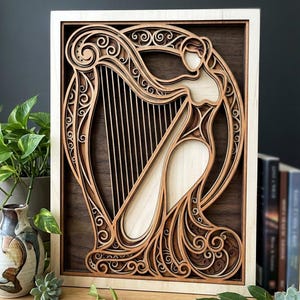 May include: A layered wooden art piece featuring a stylized harp and a female figure. The artwork is framed in light wood, with the harp and figure in a lighter wood tone against a darker wood background. Intricate scrollwork details the design.